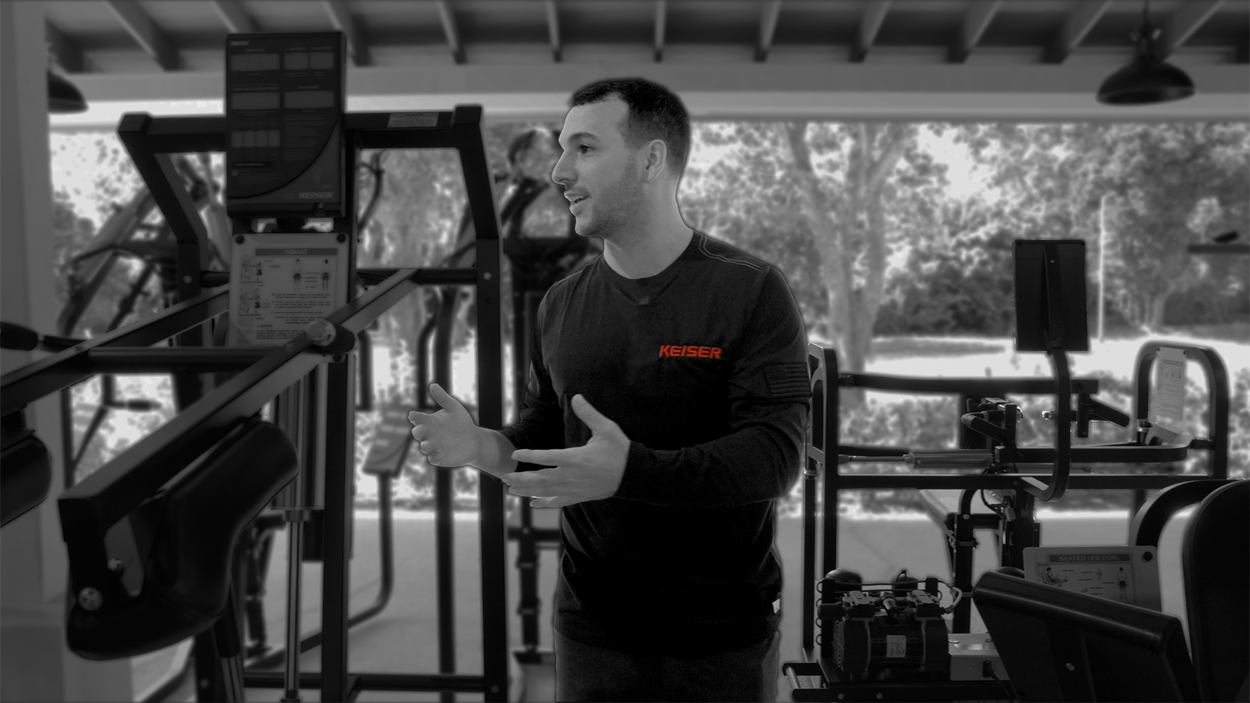 Black and white image of Keiser Human Performance and Educator Gabe standing in between a Squat Pro and Upper biaxial back